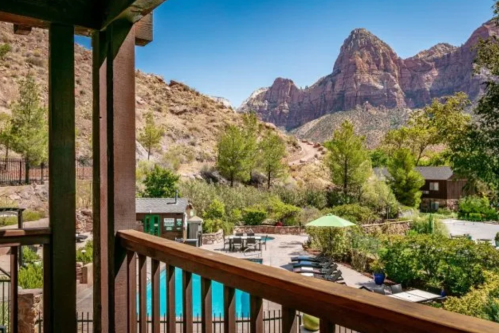 View from a balcony overlooking a pool and lush garden, framed by majestic mountains under a clear blue sky.